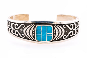 A sterling silver cuff bracelet featuring a central turquoise inlay and detailed black oxidized scrollwork, shown on a plain white background.