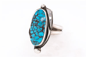 A sterling silver ring featuring a large, polished oval turquoise cabochon with a distinctive dark brown matrix pattern, set against a clean white background.