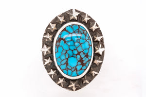 An oval turquoise ring featuring a vibrant blue-green stone with dark brown matrix veining, surrounded by a silver-toned bezel and a border of raised star details.