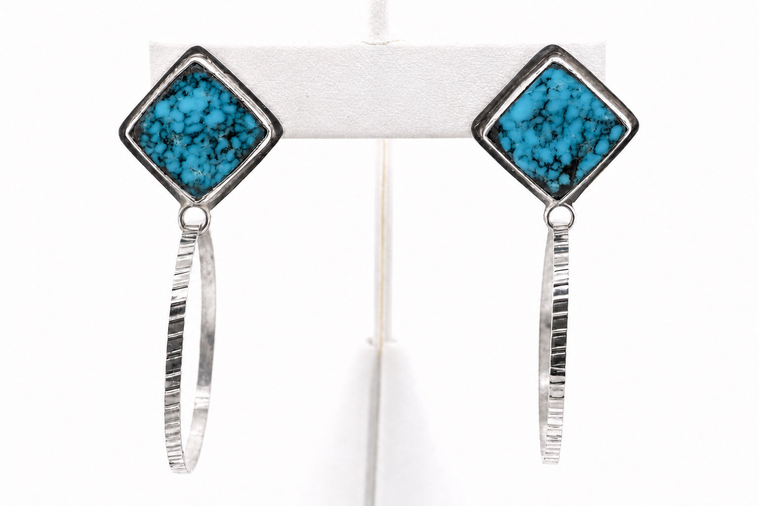 A pair of silver hoop earrings with square Egyptian turquoise stone settings, displayed on a white jewelry stand against a white background.