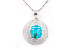 A silver pendant necklace featuring a textured circular silver frame with a central turquoise cabochon stone, suspended from a silver chain against a plain white background.